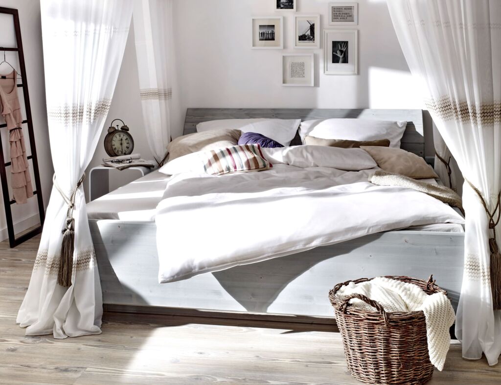 Hellgraues Holzbett von Tasso mit weißen Himmelbett-Vorhängen und einem dunkelbraunen Rattankorb auf hellem Holzboden