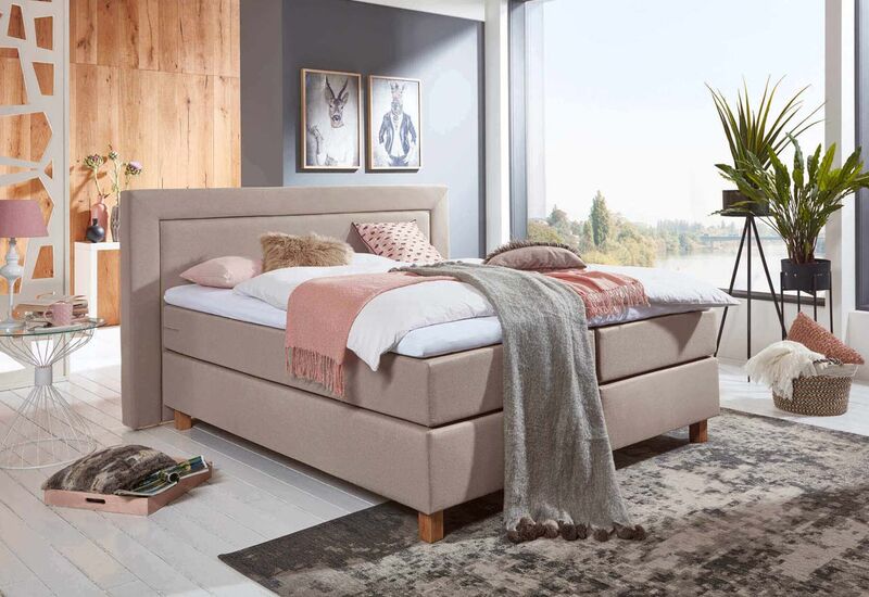 Beiges Boxspringbett, freistehend im Raum vor einer hellen Fensterfront.