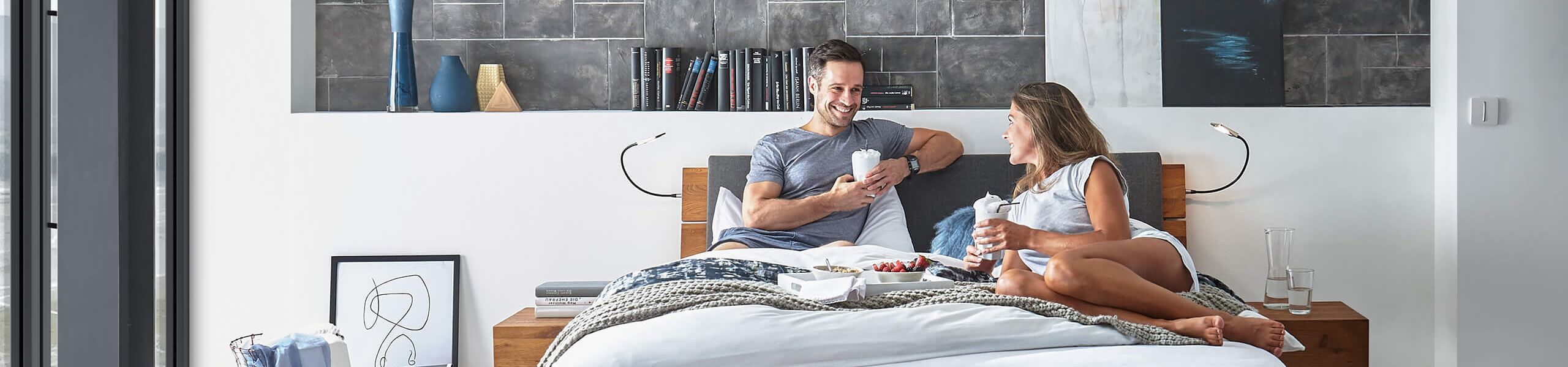 Ein Paar sitzt entspannt auf einem modernen Bett aus Wildeiche und genießt gemeinsam Frühstück im hellen Schlafzimmer.