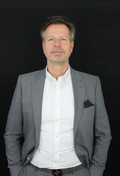 Rene Wolber steht in einem grauen Anzug und weißem Hemd vor einem schwarzen Hintergrund.