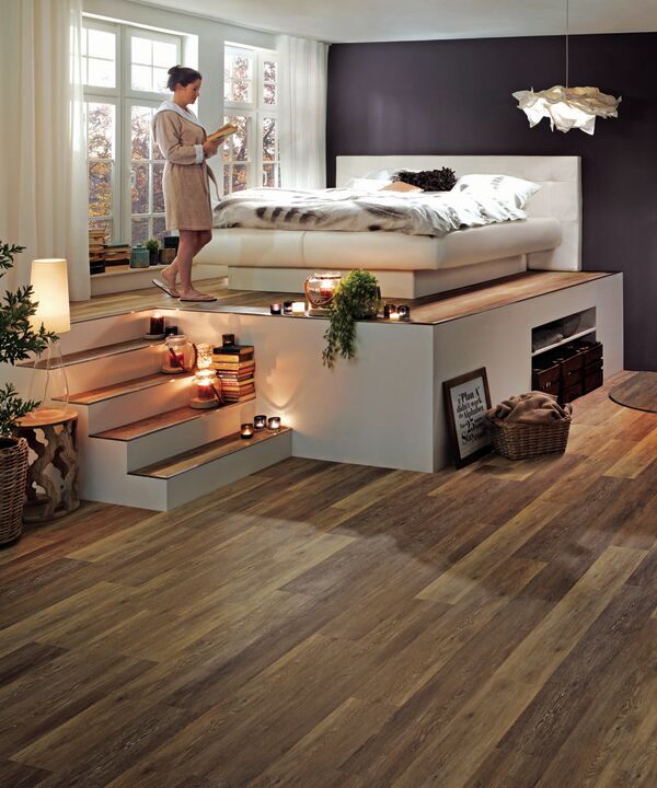 Weißes Tasso-Bett auf einer Empore in einem holzlastigen Raum mit dunkler Wand und hellen Akzenten mit weiblichem Model im Morgenmantel