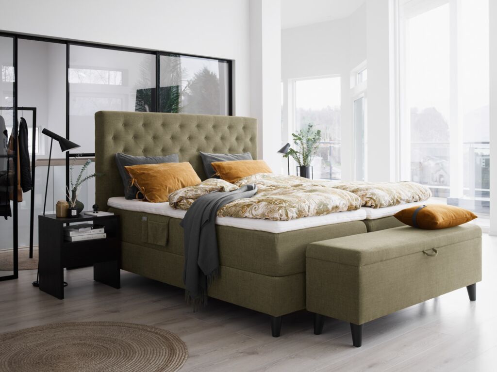 Hell-olivgrünes Boxspringbett von Svane mit farblich passender Bettwäsche und Kissen in honiggelb und grau in einem hellen Raum mit großen Fenstern und hellem Holzboden