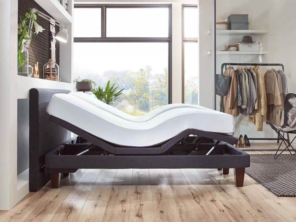 Ein verstellbares Bett in einer modernen Wohnung ist aufgerichtet, während Tageslicht durch große Fenster strömt.