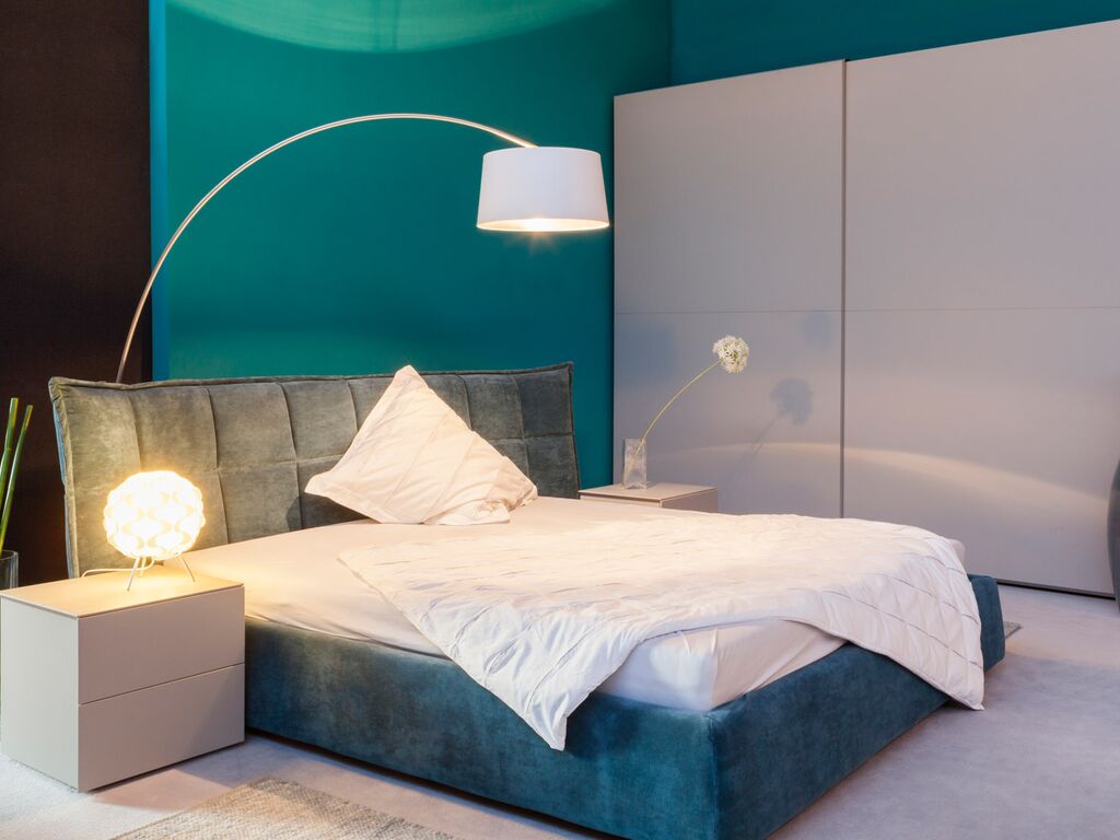 Ein blaues Stoffbett vor einer grau-türkisen Wand mit einer großen Stehlampe und grauen Möbeln im Bettenfachgeschäft Köln.