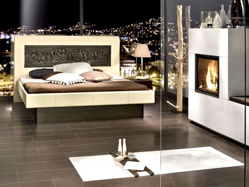 Cremefarbenes Tasso-Bett mit spiegelndem Fußteil auf dunkelbraunem Boden in einem stimmungsvollen Zimmer mit Kamin