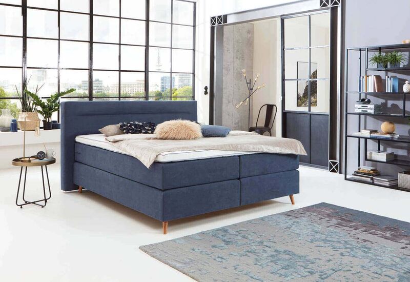Ein modernes Schlafzimmer mit einem freistehenden blauen Boxspringbett, dekoriert mit Kissen und einer gemütlichen Decke.