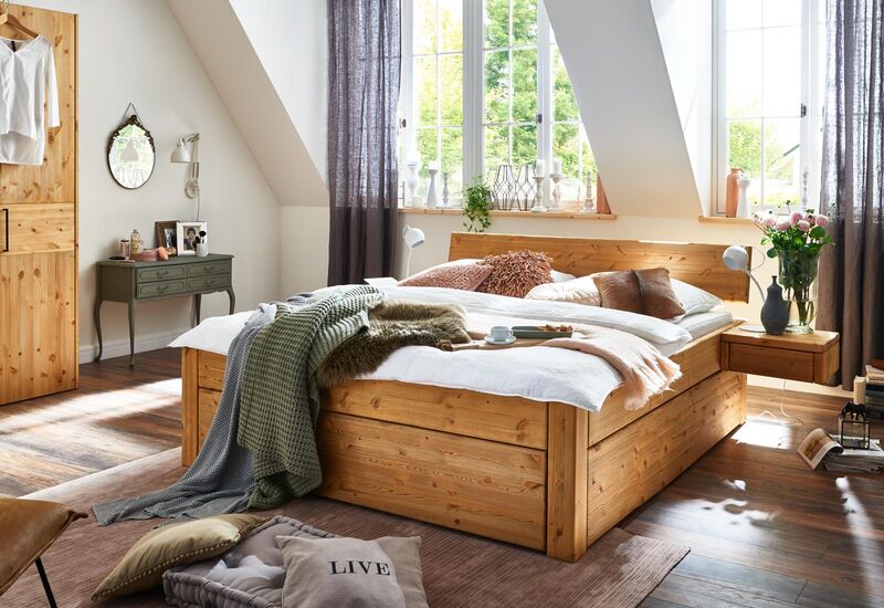 Ein Doppelbett aus massiver Kiefer steht unter einer Dachschräge mit Fenstern, durch die Sonnenlicht ins Zimmer strahlt.