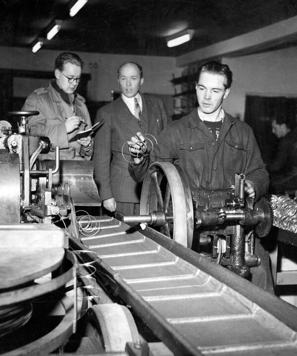 Ein Arbeiter in einer Fabrik untersucht eine Maschine, während zwei Männer seine Arbeit aufmerksam beobachten.