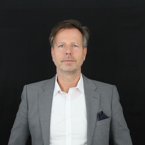 Rene Wolber steht in einem grauen Anzug und weißem Hemd vor einem schwarzen Hintergrund.