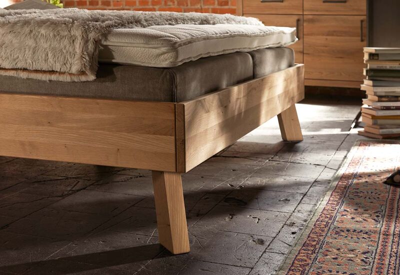 Ein Holzbett steht auf massiven, eckigen Holzfüßen auf rustikalem Boden