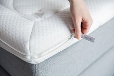 Eine Hand zieht den Reißverschluss eines weißen Matratzenaufsatzes mit dem Label SVANE auf.