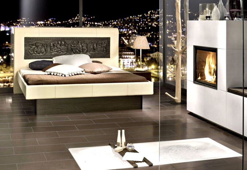 Cremefarbenes Tasso-Bett mit spiegelndem Fußteil auf dunkelbraunem Boden in einem stimmungsvollen Zimmer mit Kamin