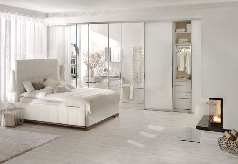 Cremeweißes Polsterbett von Tasso in einem weißen Zimmer mit großer Schrank-/Spiegelfront und bodentiefen weißen Vorhängen und Kamin