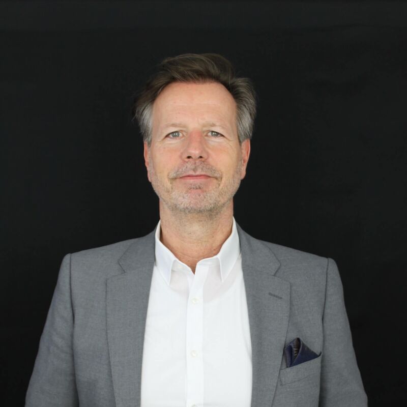 Rene Wolber steht in einem grauen Anzug und weißem Hemd vor einem schwarzen Hintergrund.