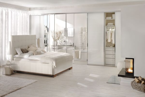 Cremeweißes Polsterbett von Tasso in einem weißen Zimmer mit großer Schrank-/Spiegelfront und bodentiefen weißen Vorhängen und Kamin