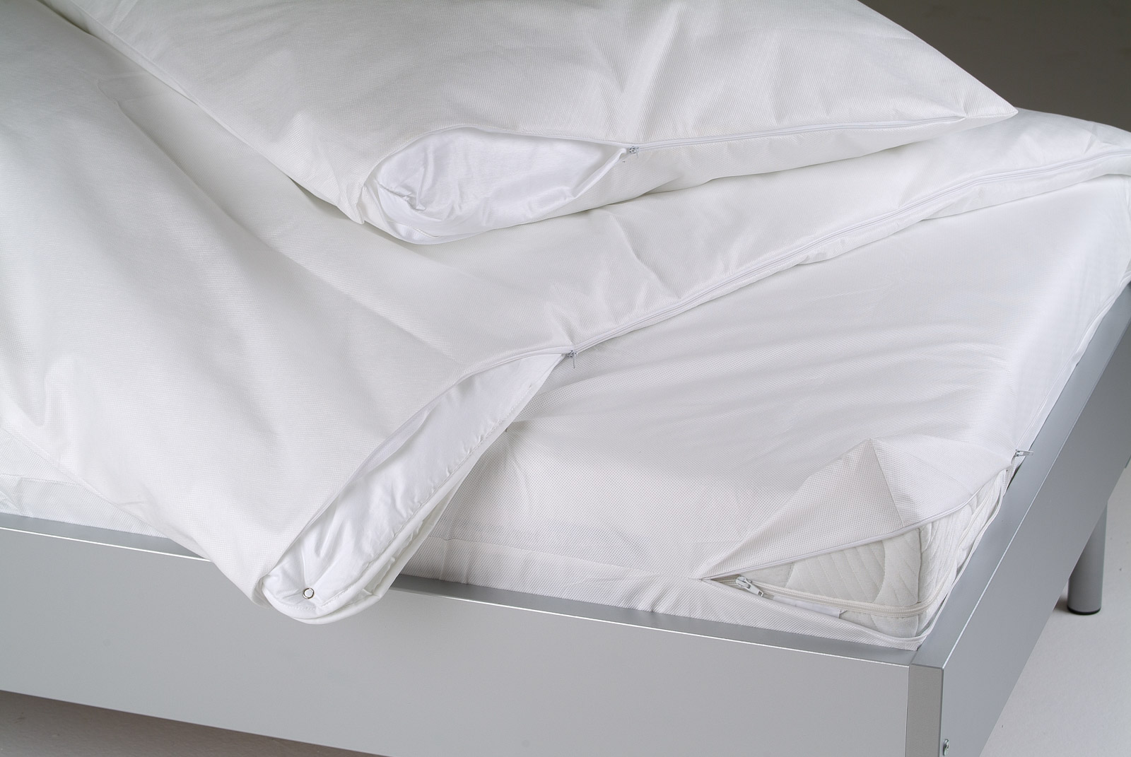 Ein frisch bezogenes Bett mit weißen Kissen und einer Decke mit Reißverschlüssen auf einem modernen, silbernen Bettgestell.