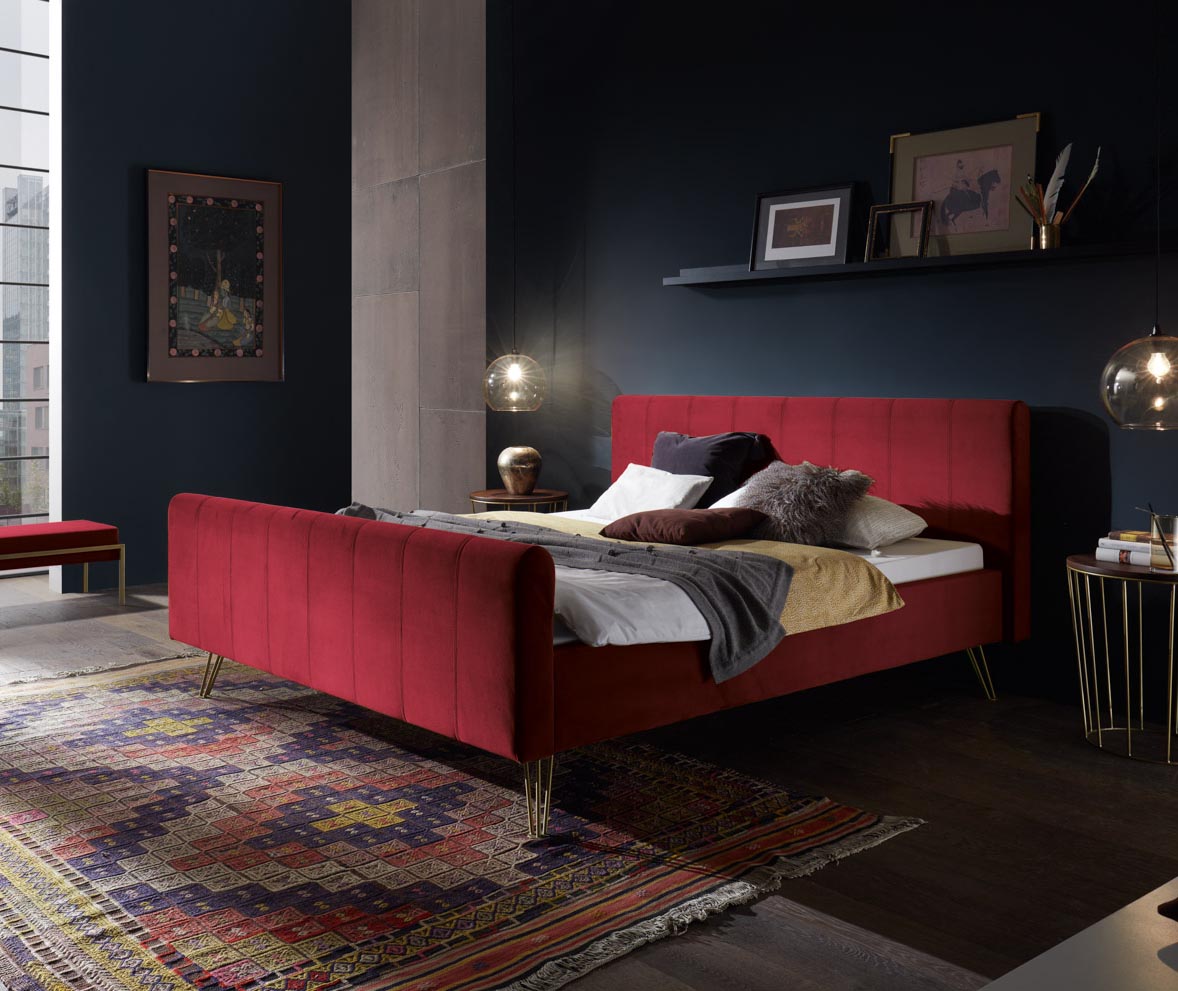 Ein sattrotes Polsterbett steht in einem stimmungsvollen Schlafzimmer mit dunklen Wänden und roten Akzenten.