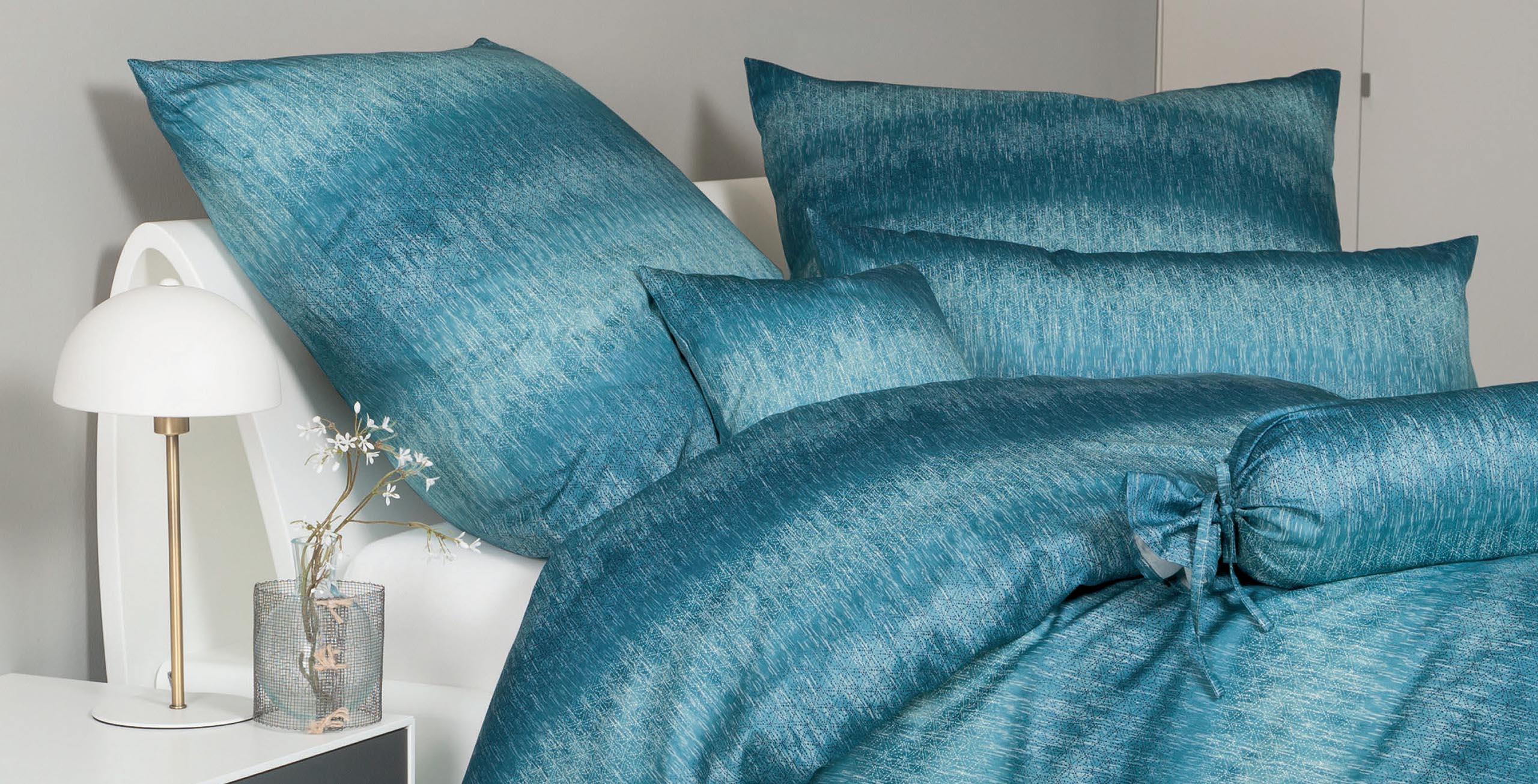 Bettwäsche in sanften Blautönen liegt ordentlich auf einem Bett, begleitet von einer modernen Tischlampe und einer Blumenvase.