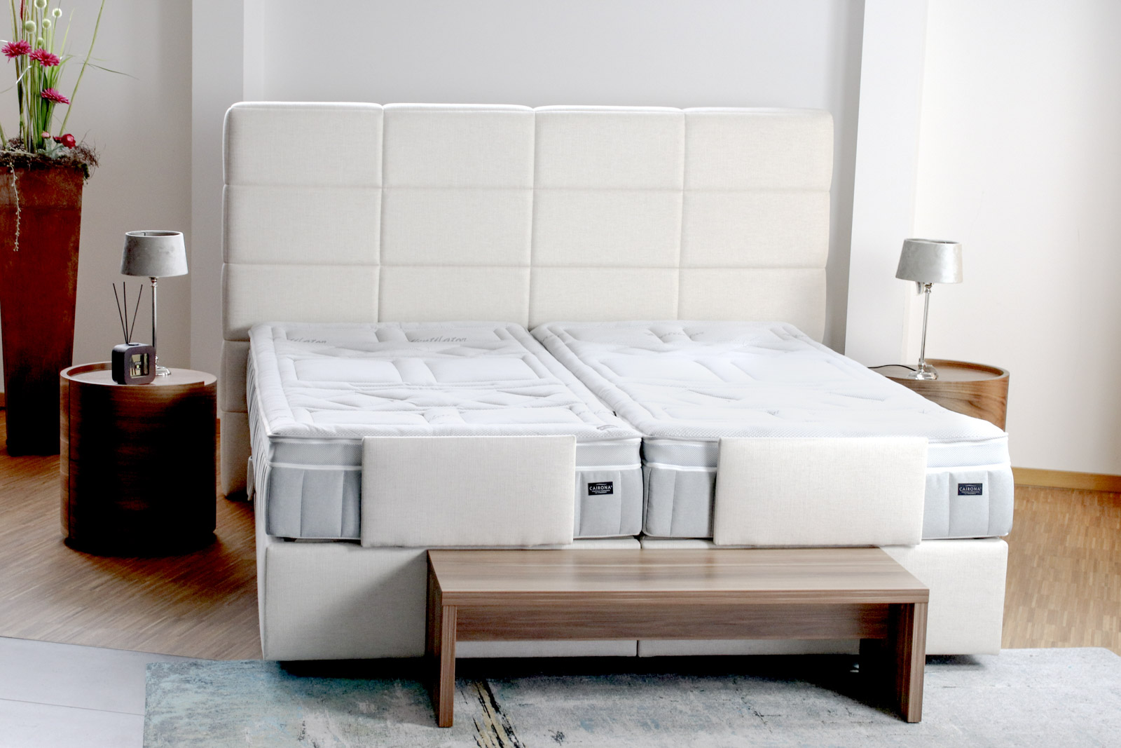 Ein Cairona-Doppelbett mit zwei Einzelmatratzen und einem eleganten Kopfteil, umgeben von stilvollen Möbeln.
