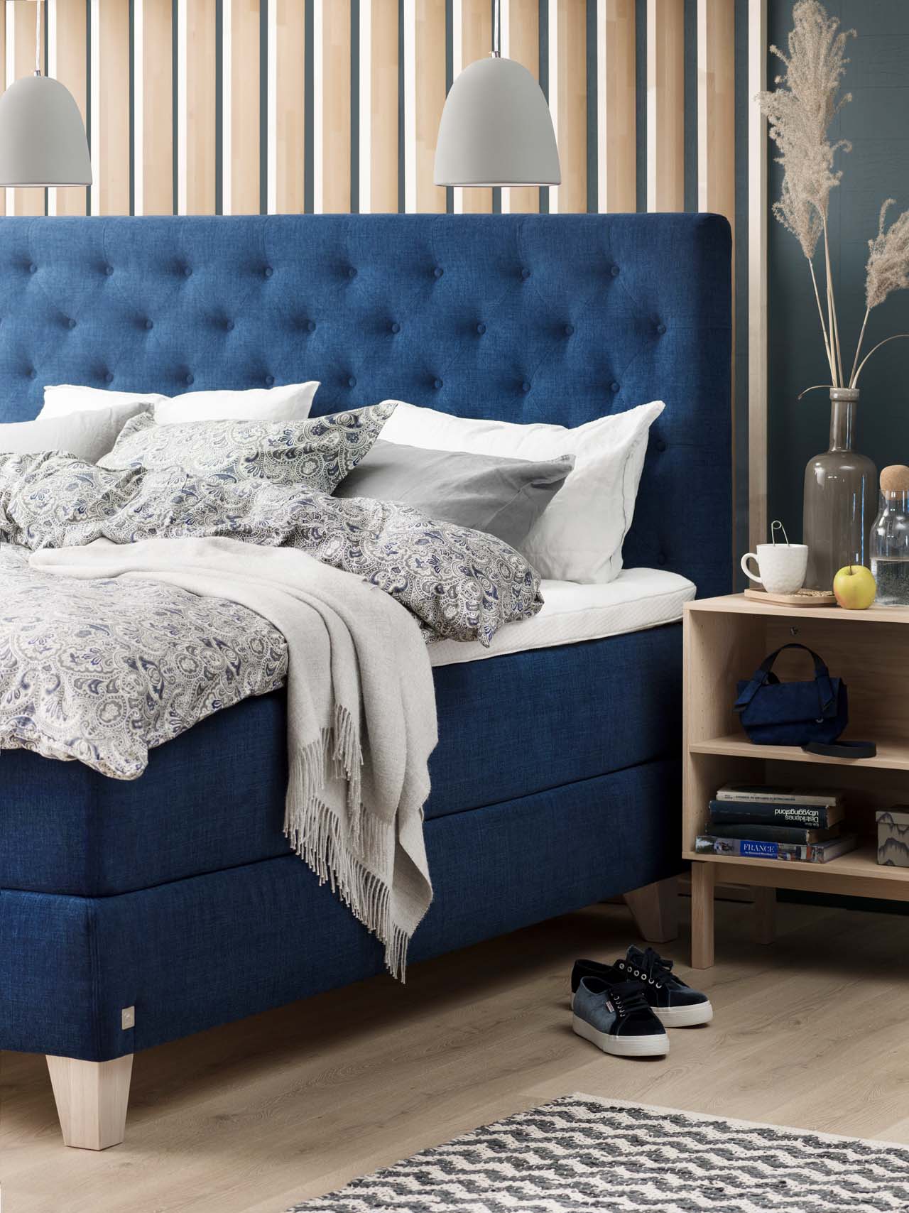 Dunkelblaues Svane-Bett im close-up mit weißen Kissen und einer grau gemusterten Decke. Daneben ein offener Nachttisch in hellem Holzton. Vor dem Bett stehen ein paar Sneaker.