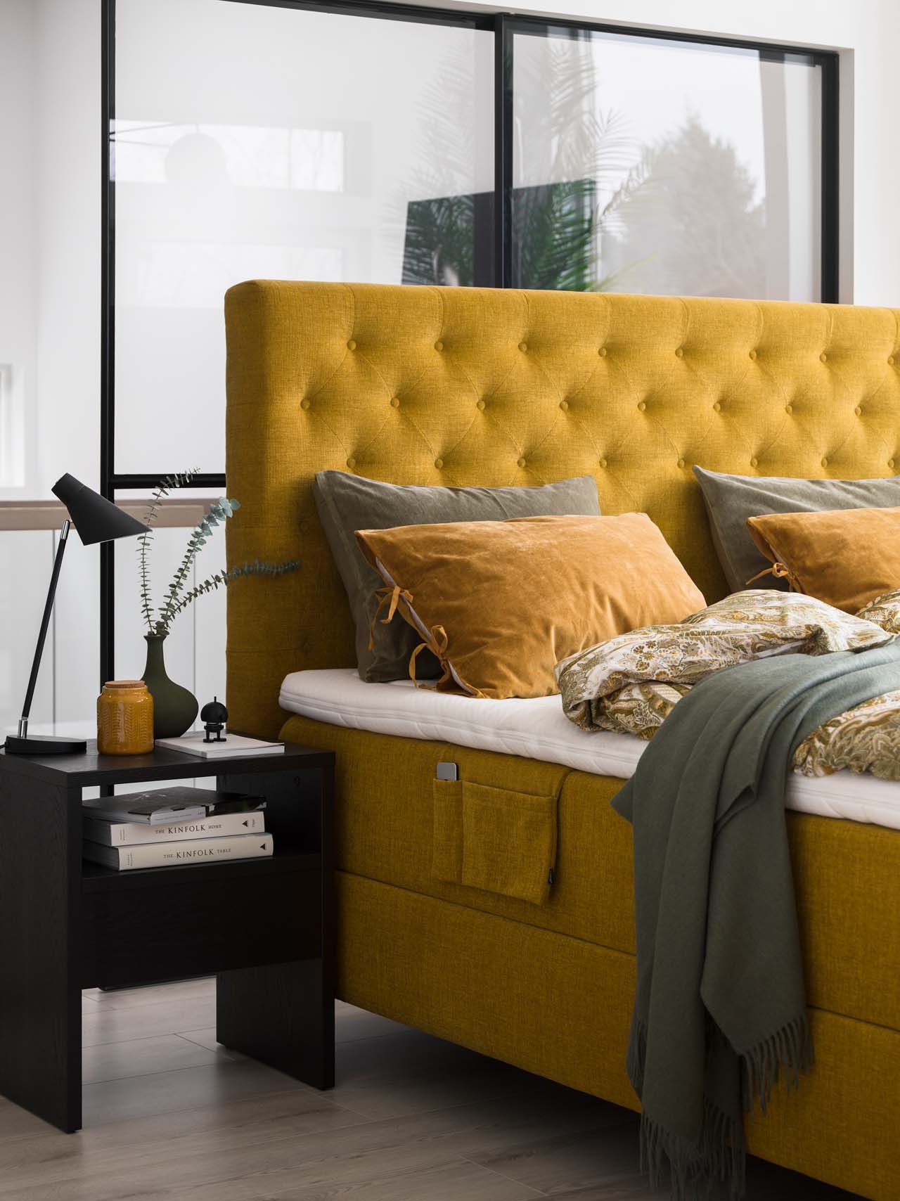 Ein senfgelb gepolstertes Bettvon Svane mit dekorativen Kissen und einem schwarzen Nachttisch.