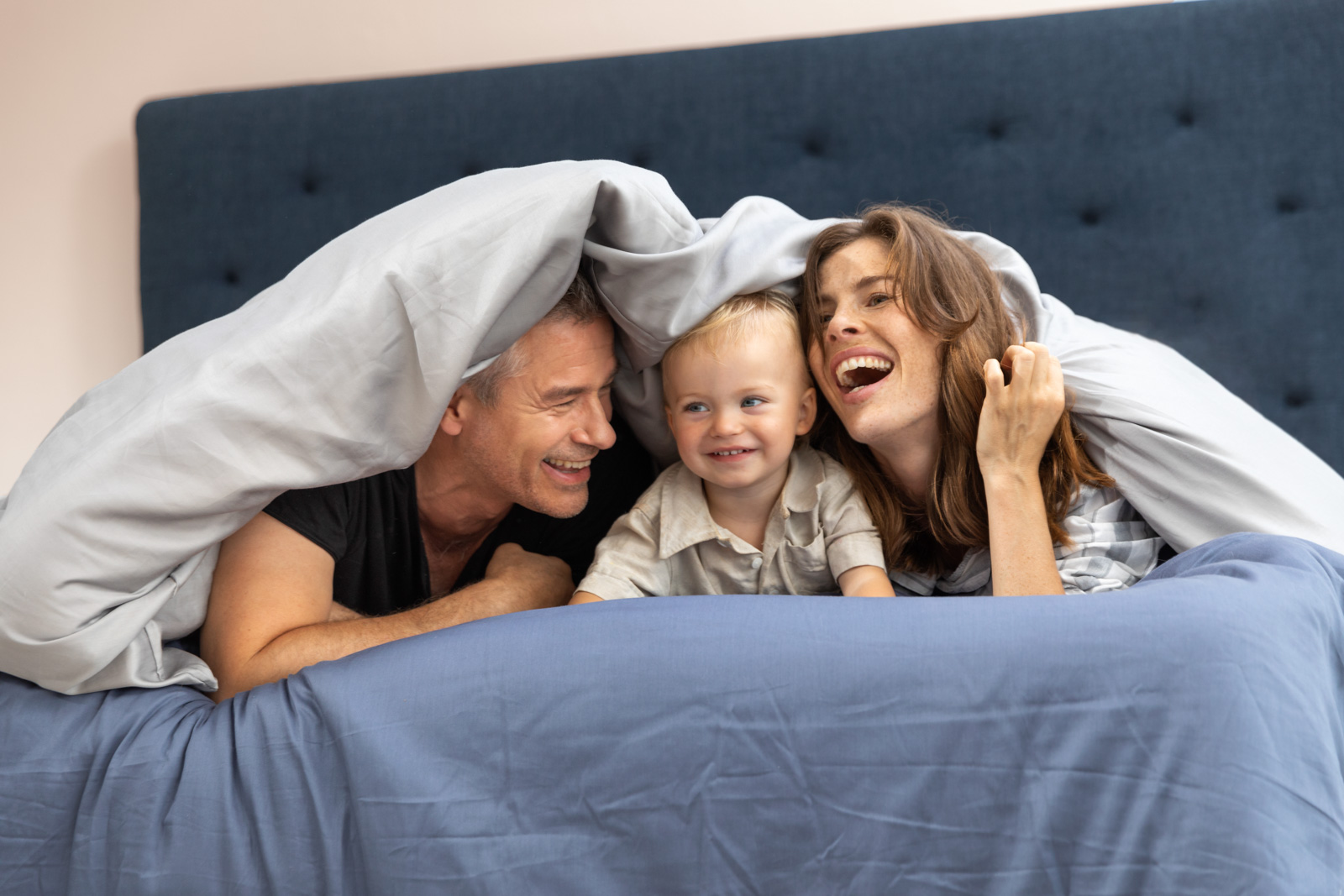 Mutter, Vater und Kind liegen in einem Svanebett mit dem Gesichtern zur Kamera, halb unter der Decke versteckt