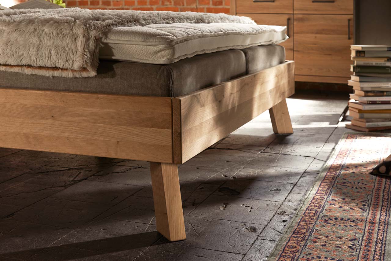 Ein Holzbett steht auf massiven, eckigen Holzfüßen auf rustikalem Boden