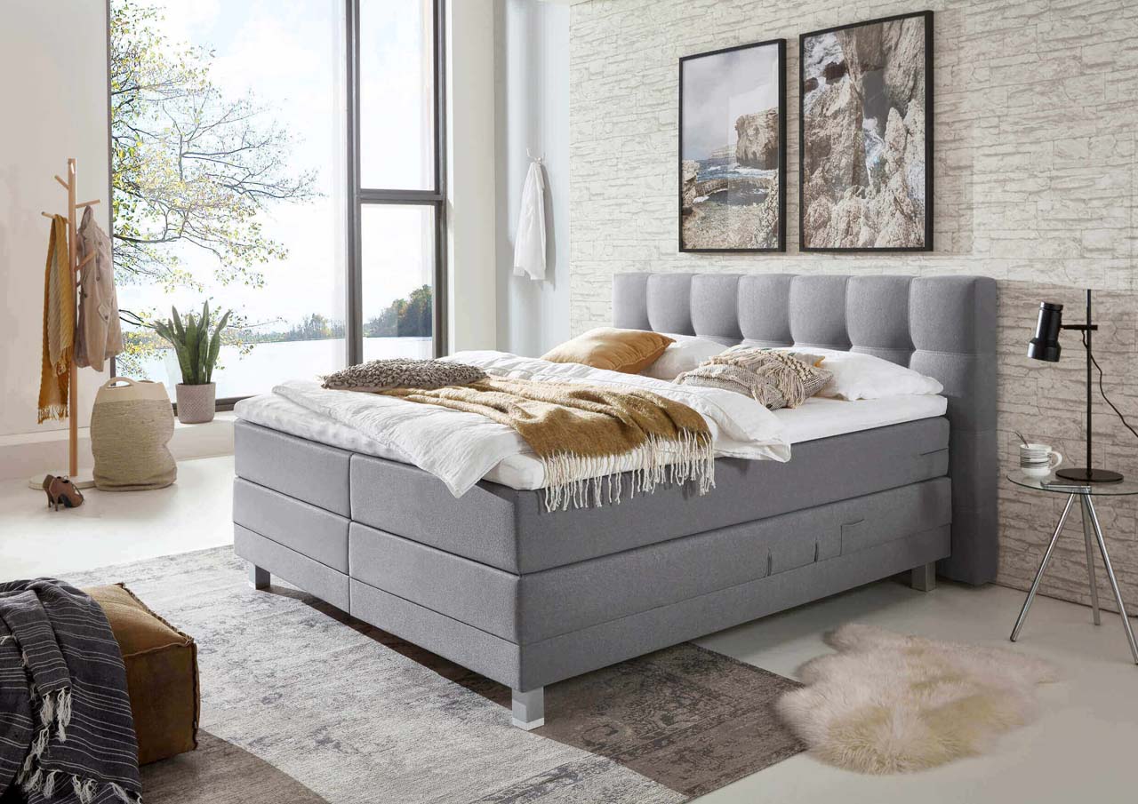 Graues Boxspringbett vor einer beigen Wand in Steinoptik in einem gemütlich eingerichteten Schlafzimmer.