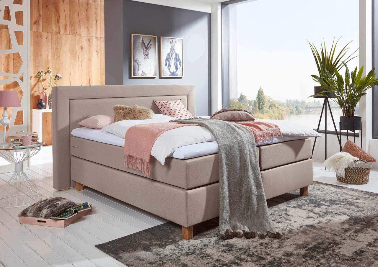 Beiges Boxspringbett, freistehend im Raum vor einer hellen Fensterfront.