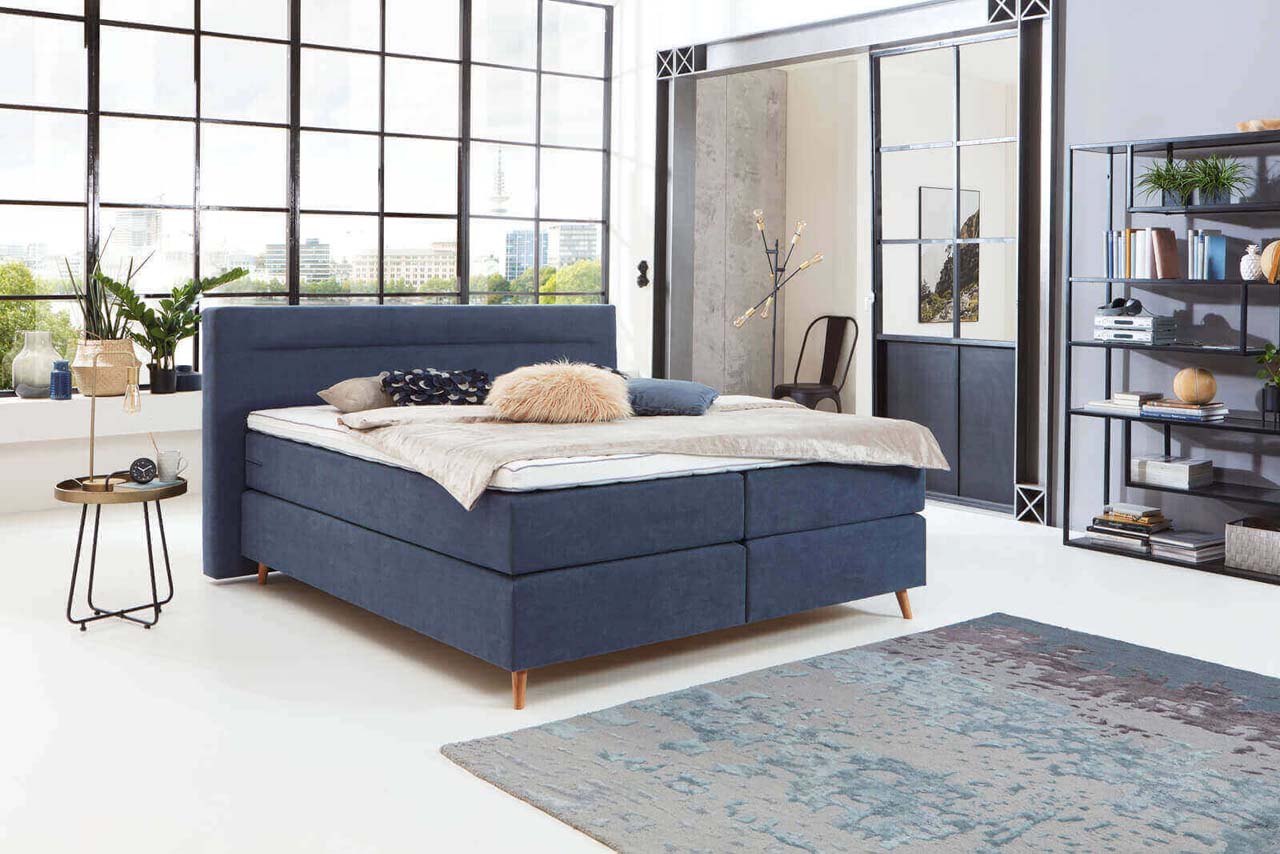 Ein modernes Schlafzimmer mit einem freistehenden blauen Boxspringbett, dekoriert mit Kissen und einer gemütlichen Decke.