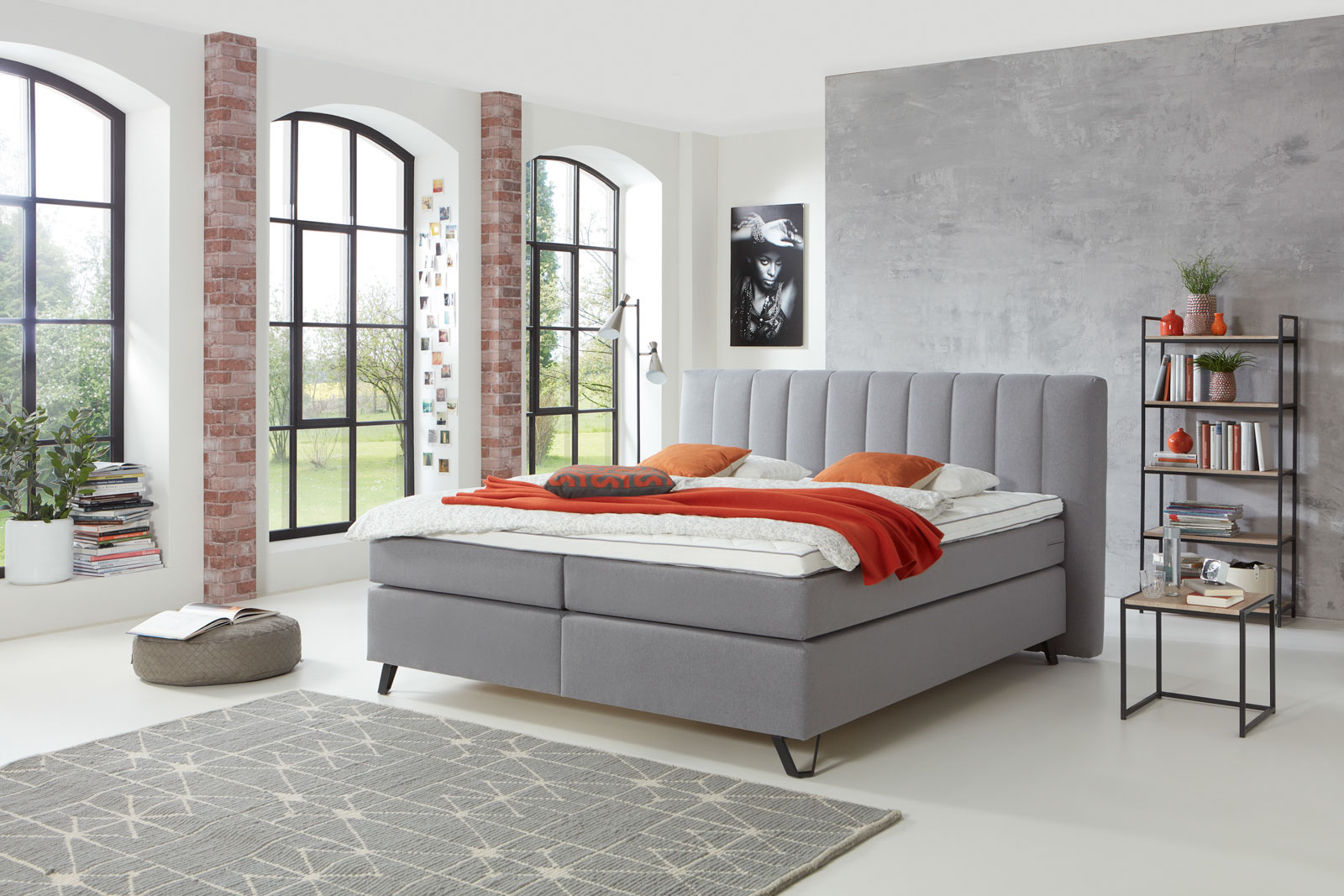 Ein stilvolles graues Boxspringbett mit roter Akzent-Decke in einem hellen Zimmer im Industrial-Stil.