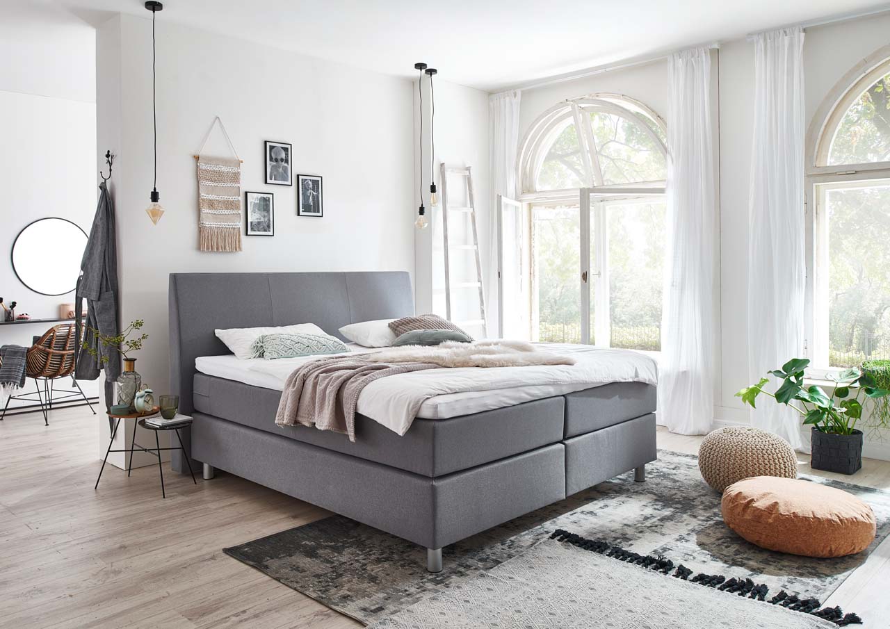 Ein modernes Schlafzimmer mit einem gemütlichen Boxspringbett, sanften Farben und großen Fenstern, die viel Licht hereinlassen.