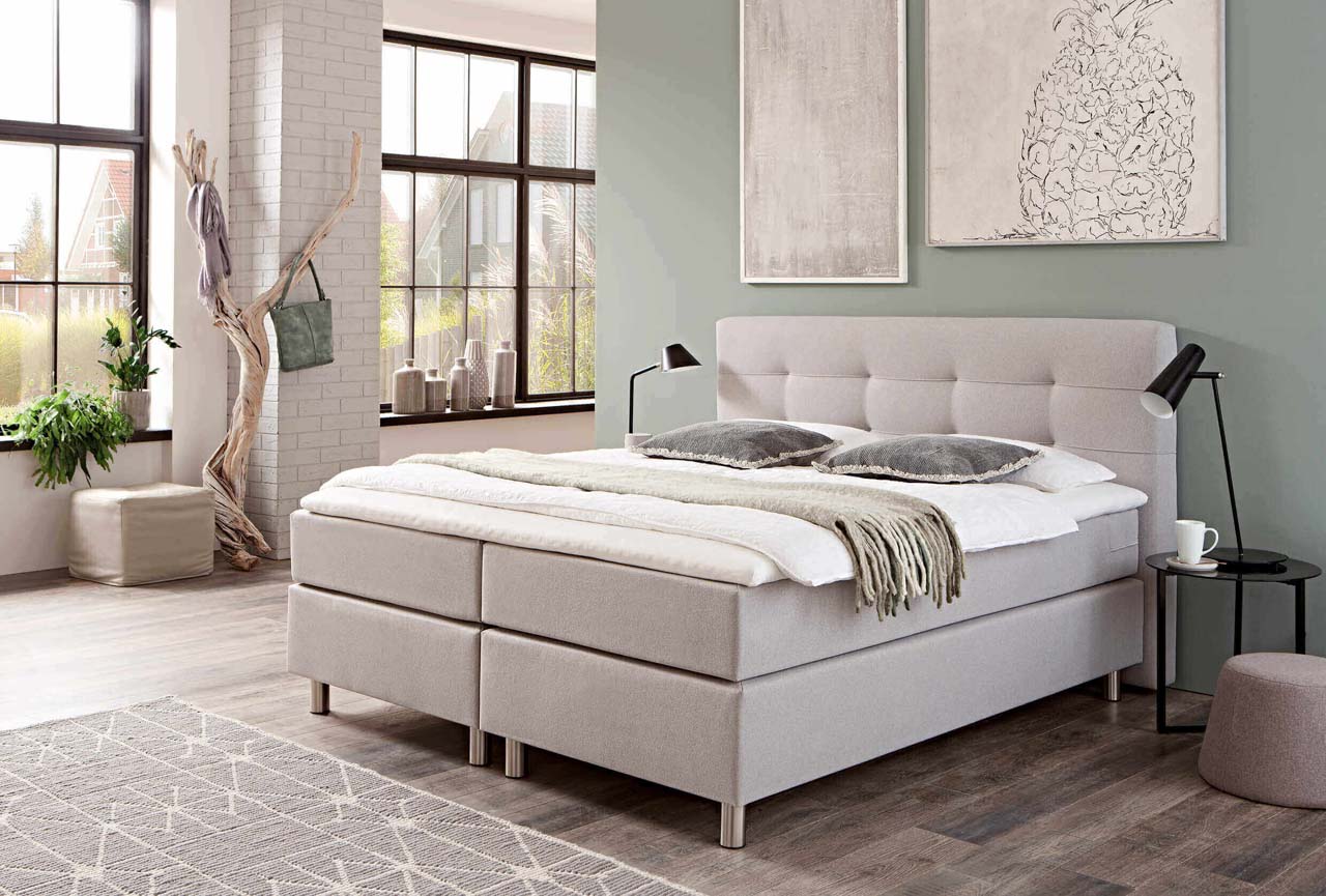 Helles Boxspringbett mit Metallfüßen vor einer salbeifarbenen Wand mit zwei großen Bildern.