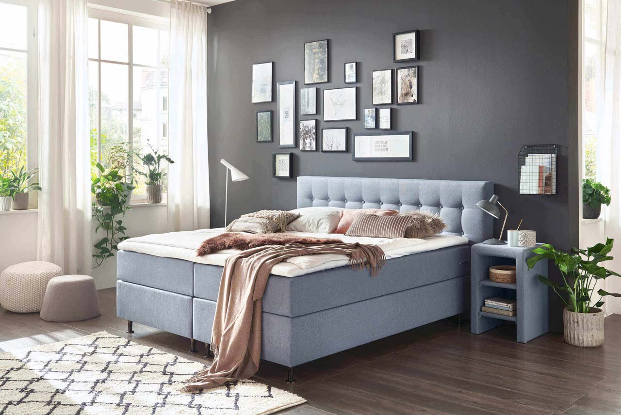 Hellgraues Boxspringbett vor einer braunen Wand mit vielen Bilderrahmen.