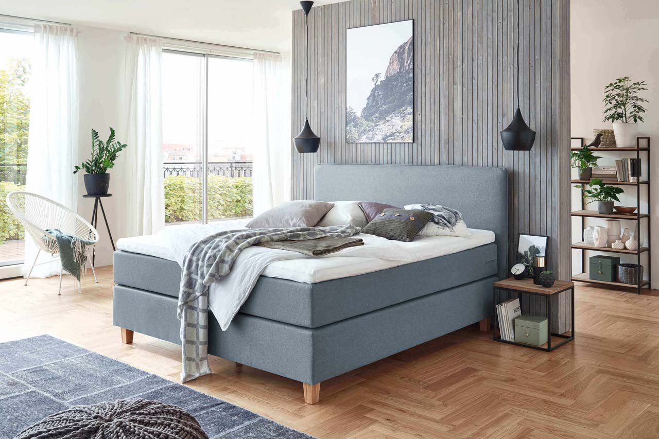 Ein stilvolles Schlafzimmer mit einem bequemen Boxspringbett, natürlichen Materialien und großen Fenstern, die Tageslicht hereinlassen.