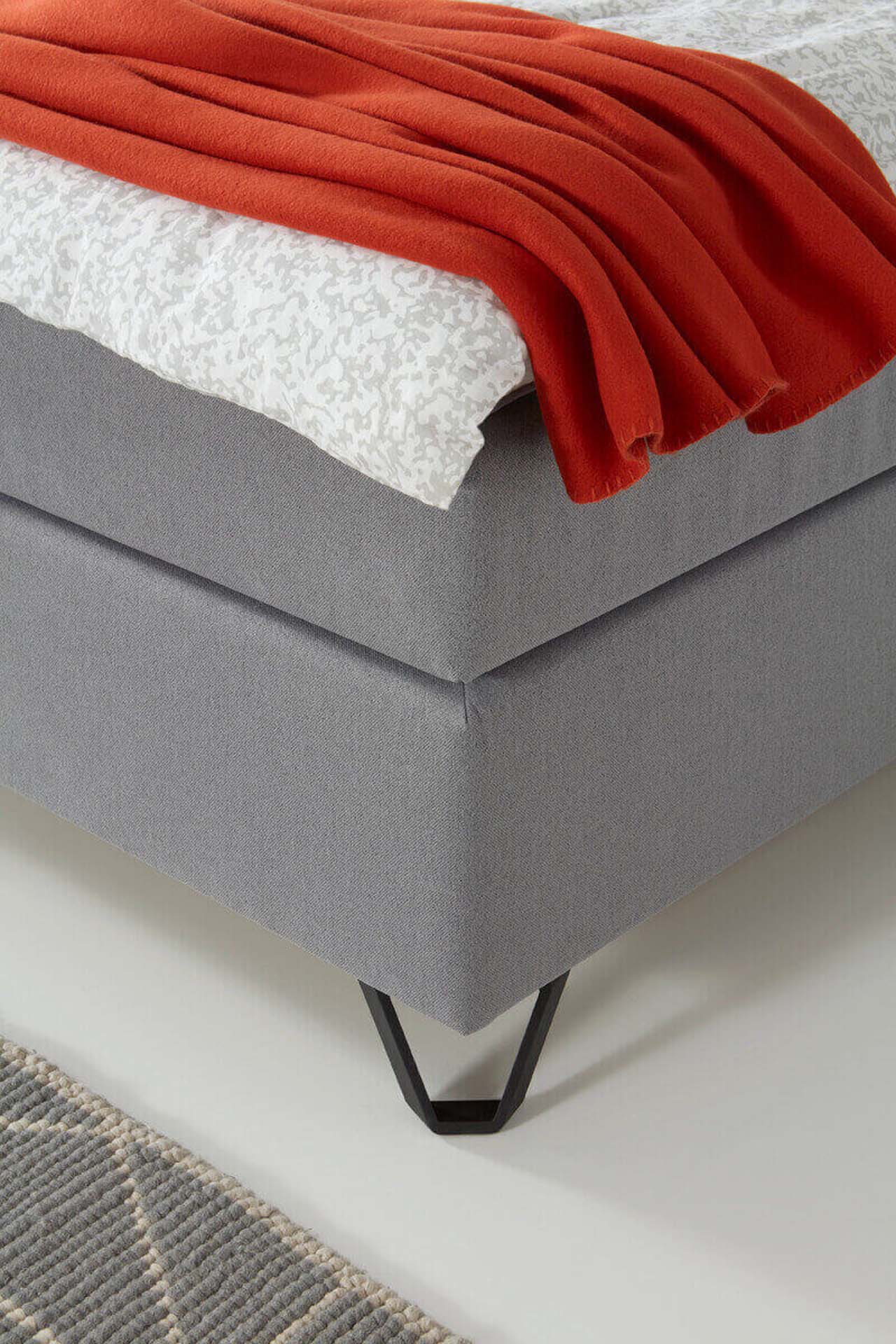 Nahaufnahme eines Fußteils eines grauen Boxspringbetts mit einer roten Decke.