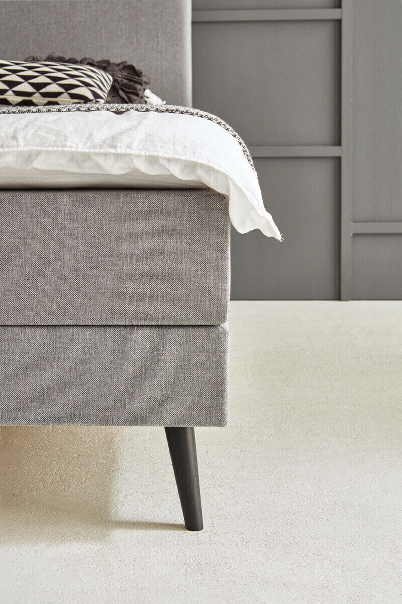 Nahaufnahme eines Fußteils eines grauen Polsterbettes mit schlankem Möbelfuß.