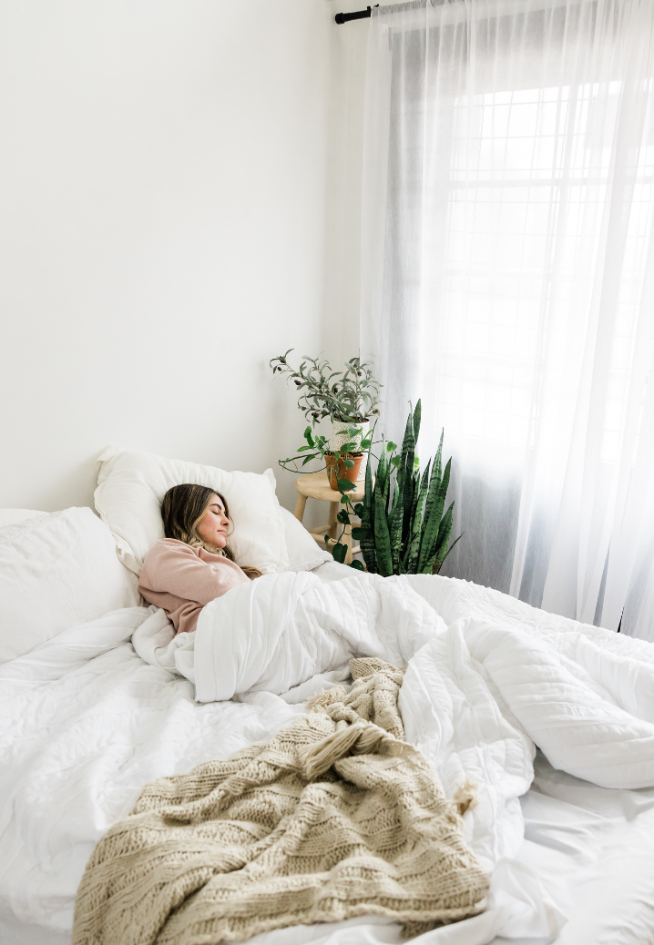 Eine Frau schläft friedlich in einem Bett mit weißer Bettdecke, umgeben von Pflanzen.