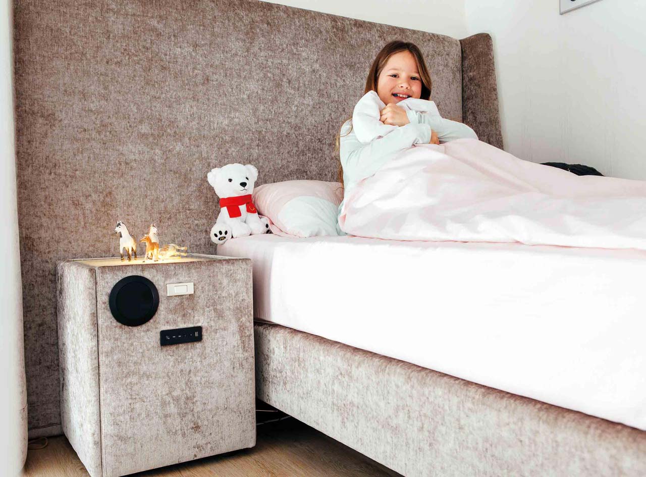 Ein lächelndes Kind sitzt in einem hellen Stoffbett mit farblich passendem Nachttisch, neben dem Mädchen ein weißer Teddy mit rotem Schal.