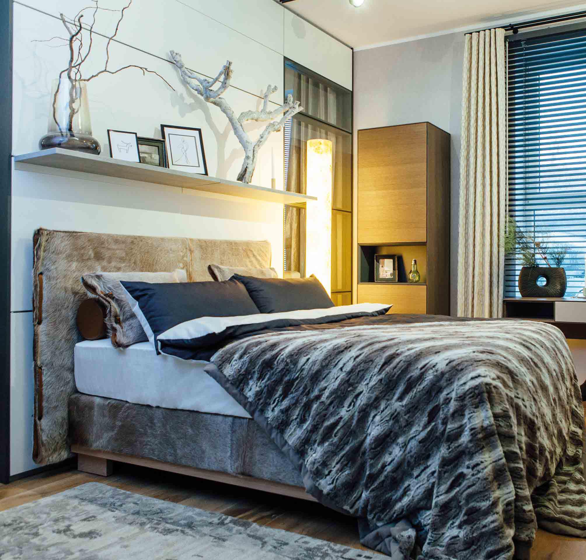 ein Bett mit Felloptik in Grau-braun-Tönen von Keno Kent in schräger Seitenansicht mit passender Decke und mehreren Kissen.